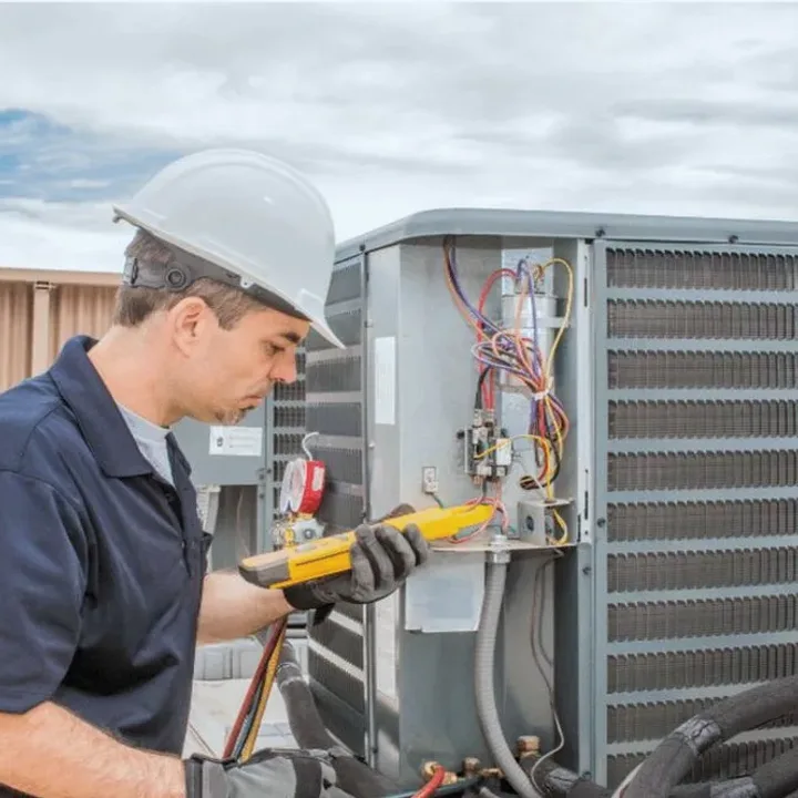HVAC technician in a hard hat testing an outdoor air conditioner with a multimeter
